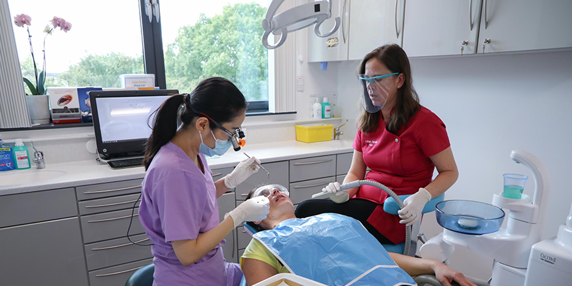 patient lying in dental chair receiving treatment from dentist is accompanied by dental nurse